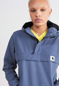 Giacca leggera a pullover di colore blu, con cappuccio, tasca frontale e fodera nera. Presenta un patch con logo sul lato sinistro.
