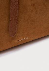 Bruine suède tas met een glad leren riem en goudkleurig, gegraveerd "Coccinelle"-logo. Zachte textuur en rechthoekige vorm.