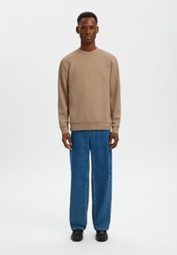 Μπεζ φούτερ με στρογγυλή λαιμόκοψη και μακριά μανίκια, σε συνδυασμό με φαρδιά μπλε τζιν παντελόνια και μαύρα παπούτσια. Απλός, καθαρός σχεδιασμός χωρίς σχέδια.