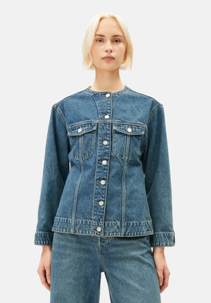 Junge Frau mit kurzen blonden Haaren trägt eine geknöpfte, langärmelige Jeansjacke und passende Jeanshose, steht vor einem schlichten Hintergrund.