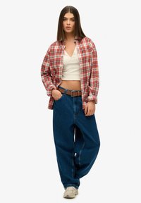 Chemise en flanelle à carreaux rouges et blancs portée sur un haut blanc à col en V, associée à un jean en denim bleu ample et des baskets beiges. Une ceinture marron met en valeur les détails.