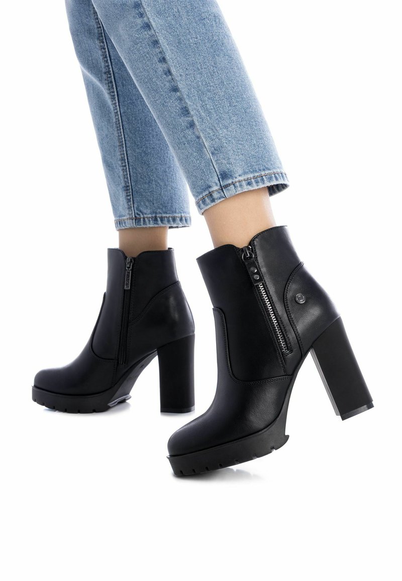 Botas de cuero negras hasta el tobillo con un alto tacón cuadrado, cremalleras laterales y suelas texturizadas, combinadas con jeans azul claro doblados por el puño.