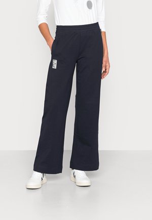 Mujer con pantalones negros de pierna ancha con un parche rectangular blanco en el muslo izquierdo y zapatillas blancas, manos en los bolsillos.