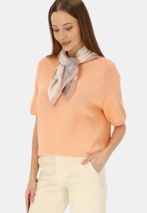 Kobieta ubrana w brzoskwiniowy sweter z krótkim rękawem, kremowe spodnie i lekki wzorzysty szal zawiązany wokół szyi, stojąca z jedną ręką w kieszeni.