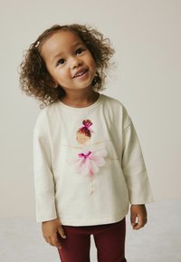 Lächelndes junges Mädchen mit lockigem Haar, das ein cremefarbenes Langarmshirt mit Ballettdesign und dunkelroten Hosen trägt, steht vor einem einfarbigen Hintergrund.