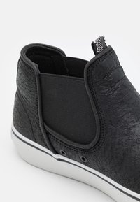 Chaussure noire texturée à enfiler avec des panneaux élastiques latéraux, une languette de maintien et une semelle en caoutchouc blanche avec une bande noire, montrée en gros plan.