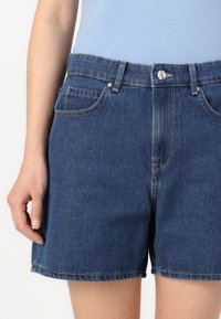 Denim shorts in dark blue, featuring a straight cut, five pockets, and silver button closure. The fabric has a smooth texture with visible stitching.