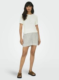 Vit, kortärmad stickad topp i kombination med ljusgrå rutiga shorts. Fötterna i bruna sandaler med korsade remmar, stående mot en enfärgad bakgrund.