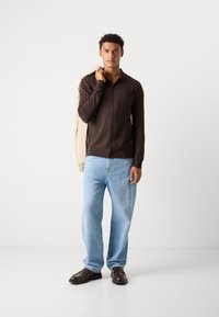 Bruine overhem shirt met lange mouwen en een kraag, gedragen met lichtblauwe relaxte spijkerbroek en donkere loafers, met een beige jas in de hand.