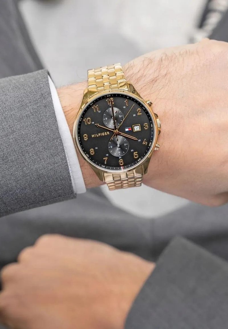 Auksinės spalvos chronografas su juodu ciferblatu, trimis subdailiais, datos rodymu ir auksinės spalvos metaline apyranke, rodoma ant riešo dėvint pilką kostiumą.