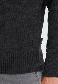 Close-up van een donkergrijze gebreide trui manchet en zoom met een klein stoffen merklabel, gedragen over lichtgrijze pantalons.