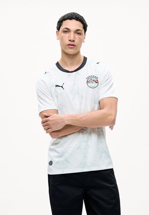 Jaunas vyras trumpais garbanotais plaukais, dėvintis baltą sportinį marškinėlį su juodu apykakliu ir Egipto futbolo komandos emblemą, sukryžiuotomis rankomis.