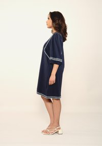 Dunkelblaues Kleid mit kurzen Puffärmeln, verziert mit weißer Stickerei entlang des Ausschnitts und Saums, kombiniert mit weißen, strappy Blockabsatzsandalen.