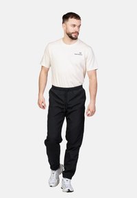 Zwarte elastische broek met een trekkoord, gecombineerd met een lichtbeige t-shirt met ronde hals waarop een klein logo op de borst staat.