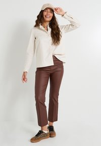 Chemise beige clair à boutons avec col volanté, pantalon en simili cuir marron et baskets imprimé léopard, portant une casquette claire avec inscription.