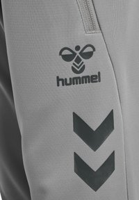 Zbliżenie na szarą tkaninę z czarnym logo hummel oraz dwoma skierowanymi w dół paskami w kształcie szewronów obok kieszeni zamykanej na zamek.