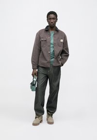 Šedá džínová bunda s náprsními kapsami, zelená košile, tmavé džíny, béžové boty a malá zelená kabelka. Jednoduchý, moderní design.