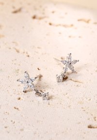 Orecchini a forma di stella argentata con cinque cristalli trasparenti a taglio marquise, con un'illuminazione pendente di un cristallo più piccolo, esposti su una superficie testurizzata.