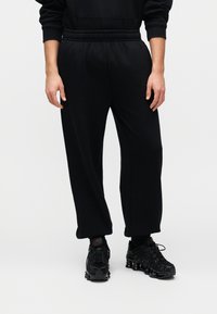 Μαύρα βαμβακερά παντελόνια jogger με ελαστική μέση, άνετη γραμμή και τσέπες στο πλάι, συνδυασμένα με μαύρα αθλητικά παπούτσια με υφασμένα πέλματα.