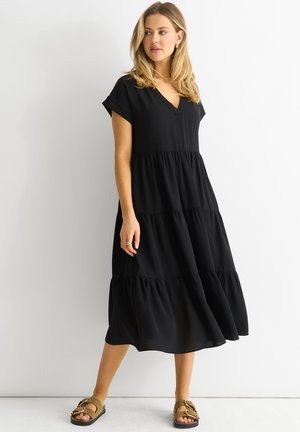 Schwarzes, gestuftes Kleid mit V-Ausschnitt, kurzen Ärmeln und lockerer Passform. Hergestellt aus leichtem Stoff, mit fließender Silhouette und knöchellang.