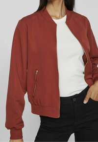 Braune Bomberjacke mit rundem Kragen, Reißverschluss und elastischen Bündchen. Verfügt über eine seitliche Reißverschlusstasche und eine glatte Textur.