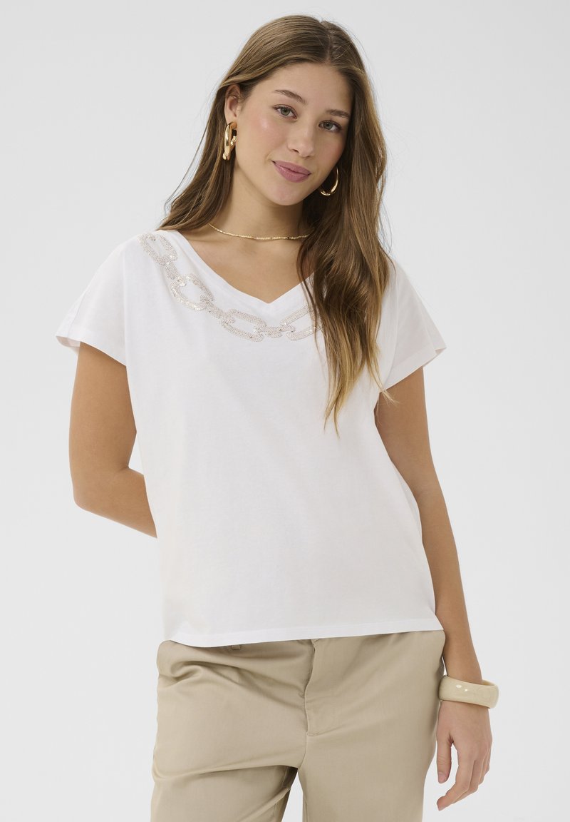 Jeune femme aux longs cheveux bruns portant un haut blanc à col en V avec un détail chaîne, un pantalon beige, des boucles d'oreilles créoles dorées et un bracelet beige.