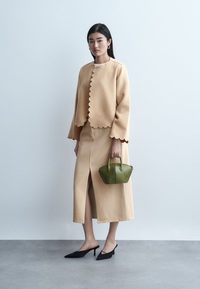 Vrouw draagt een beige jasje met geschulpte randen en een bijpassende rok met een vooraan gespleten zoom, houdt een kleine groene handtas vast en staat op een grijze vloer tegen een witte muur.