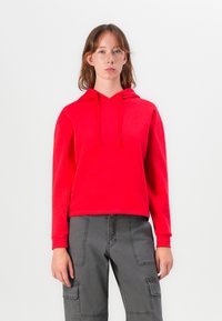 Sarkans džemperis ar kapuci, ar vaļēju piegriezumu un garām piedurknēm. Saskaņots ar pelēkiem pielaikojošiem biksēm, kurām ir kabatas un šuves.