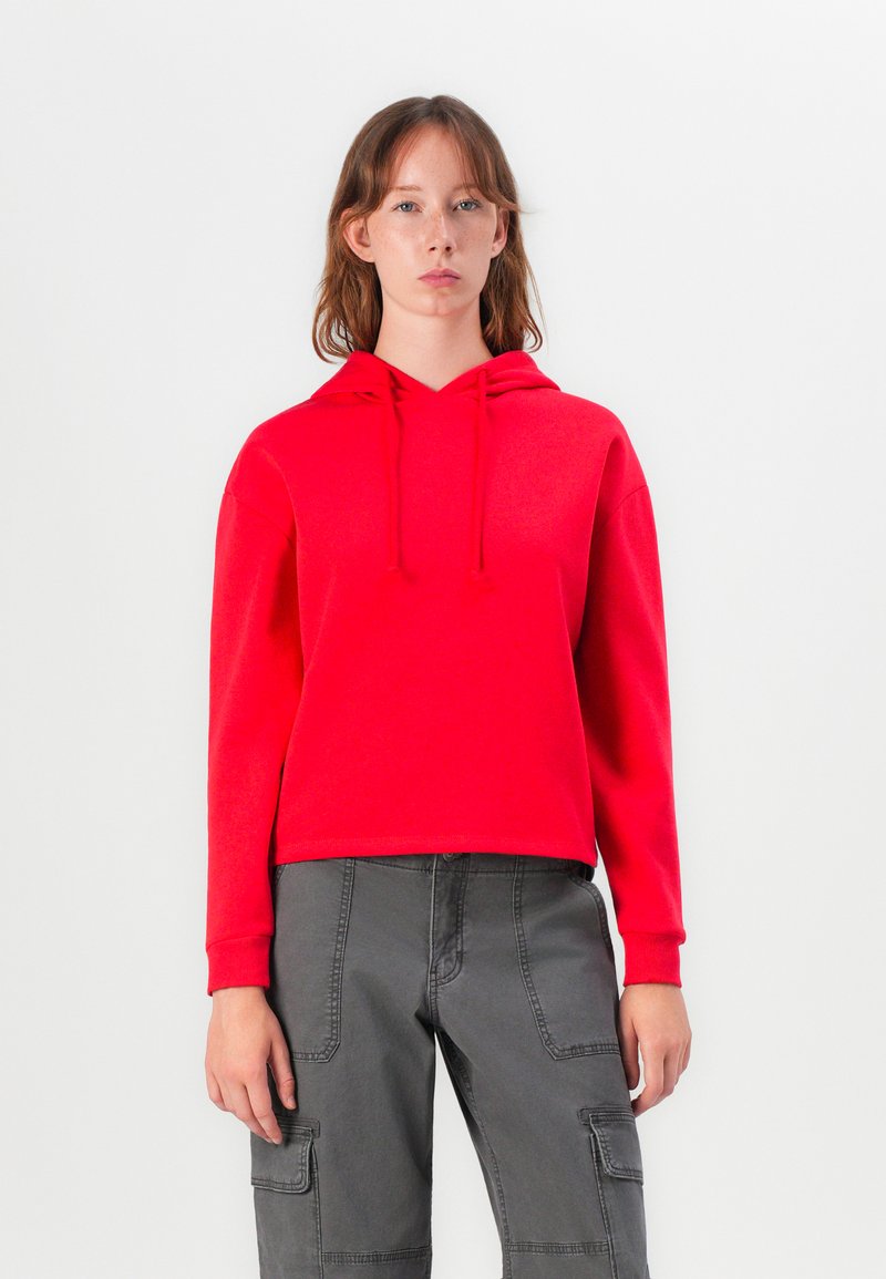 Κόκκινο φούτερ με κορδόνι, χαρακτηριστικό χαλαρού φίτινγκ και μακριά μανίκια. Συνδυάζεται με γκρίζες cargo παντελόνες που διαθέτουν τσέπες με καπάκια και ραφές.