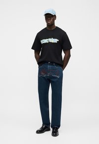 Μαύρο μπλουζάκι με το κείμενο "Icecream" σε μπλε και κίτρινες αποχρώσεις, συνδυασμένο με σκούρο μπλε τζιν με κόκκινες και λευκές λεπτομέρειες. Απαλό μπλε καπέλο, μαύρα παπούτσια.