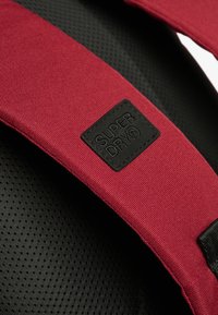 Correa de hombro de tela roja con respaldo de malla acolchada negra y un parche de logo texturizado que dice "SUPERDRY". Vista en primer plano de los materiales.
