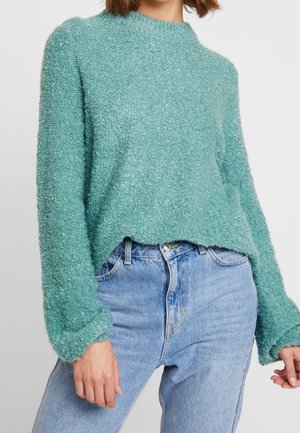Personne portant un pull texturé bleu sarcelle clair à manches longues associé à un jean taille haute bleu clair sur un fond uni.