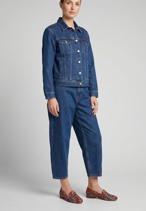 Žena stojící v modré džínové bundě a odpovídajících džínách s barevnými mokasíny s květinovým vzorem na jednobarevném pozadí.