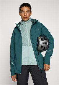 Jachetă waterproof de culoare verde cu un strat interior de un albastru deschis, având un guler înalt și accente reflectorizante, ținută alături de o cască neagră.