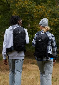 Svart ryggsekker med logodetaljer, justerbare stroppar og ekstra oppbevaring; båret av to individer i friluftsklær mot en naturskjønn bakgrunn.