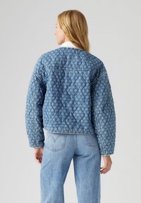Quiltana jeans jakna v svetlo modri barvi, katere značilnost je strukturiran valovit vzorec, skrajšana zasnova in kontrastno šivanje. V kombinaciji z svetlo modrimi kavbojkami.