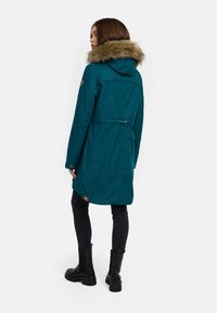 Parka teal con cappuccio in pelliccia sintetica, vita aderente e dettagli con bottoni. Maniche lunghe, spacco sul retro e orlo inferiore che arriva a metà coscia. Jeans neri e stivali.