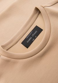 T-shirt beige à col rond avec étiquette noire affichant "OMBRE EXIMIO", "QUALITÉ PREMIUM", et la taille "M" à l'intérieur du col.