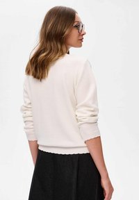 Witte gebreide trui met lange mouwen, gegolfde zoom en ribgebreide manchetten, gedragen met een donkere rok. De achterkant toont een gladde textuur.