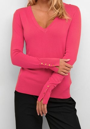 Donna che indossa un maglione a V rosa brillante con bottoni dorati sulle maniche lunghe, abbinato a pantaloni neri, in piedi davanti a uno sfondo neutro.