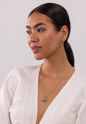 Silberne Halskette mit einem Sonnenstrahlen-Design, einem zentralen Stein und einer feinen Gliederkette. Das Modell trägt ein weißes Leinentop.