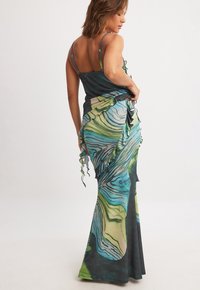 Maxi dress with adjustable spaghetti straps, featuring a wave-like green and blue print, ruffles along the side, made of soft fabric.