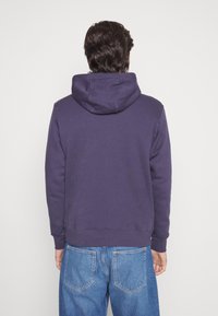 Purple hoodie with a drawstring hood, ribbed cuffs, and back hem. Paired with light blue jeans. The fabric appears soft and textured.