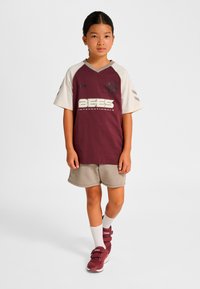 Vinröd och krämfärgad sporttröja med korta ärmar, logotyp och texten "BEES"; beigea shorts. Skor: vinröda höga sneakers med remmar.