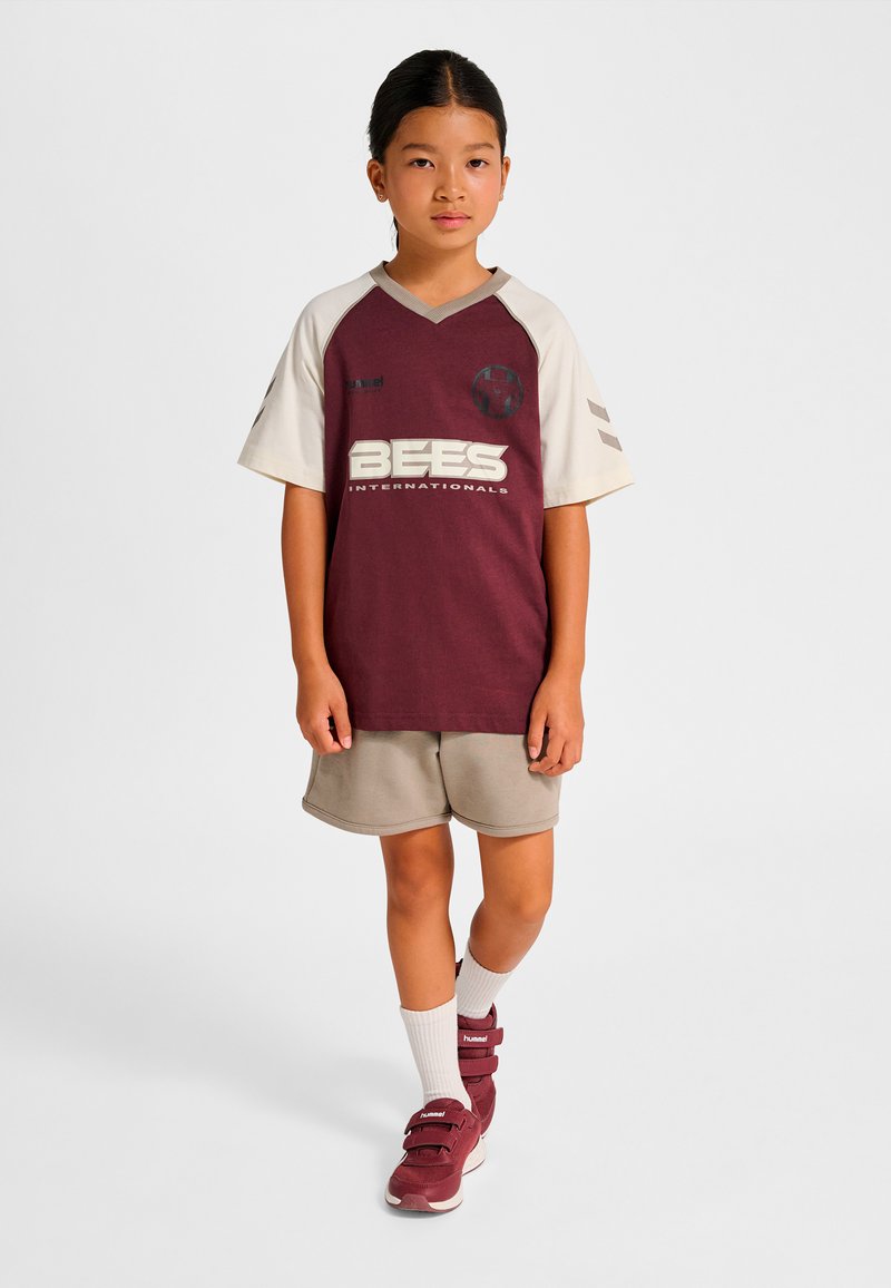 Vinröd och krämfärgad sporttröja med korta ärmar, logotyp och texten "BEES"; beigea shorts. Skor: vinröda höga sneakers med remmar.