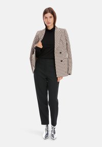 Doppeltläufiger Blazer in einem geometrischen Muster aus Schwarz und Beige. Darunter ein schwarzer Rollkragenpullover, kombiniert mit schwarzen Hosen und silbernen Ankle-Boots.
