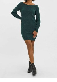 Stickad klänning i mörkgrön med båtkrage, långa ärmar och slim fit, i kombination med svarta ankelskor. Slät textur och minimalistisk design.