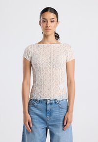 Nainen, jolla on tumma hiukset sidottuna taakse, pukee yllään valkoisen lyhythihaisen pitsipaidan ja siniset farkut, seisoo yksinkertaista taustaa vasten.