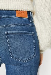 Blå denim jeans med en tætsiddende silhuet, der har et lædermærke med "ONLY" og design med en enkelt baglomme. Teksturen ser glat ud.