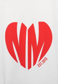 Γραφικό κόκκινης καρδιάς με στυλιζαρισμένο "NM" στο κέντρο, πάνω σε λευκό βαμβακερό t-shirt. Διαθέτει το κείμενο "EST. 2013" σε μικρό γκρι χρώμα στο κάτω δεξί μέρος.